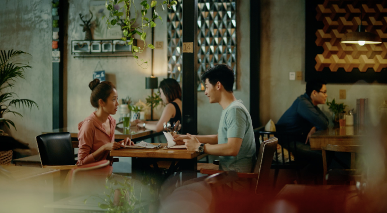 Two people sit across from each other at a table in a cozy cafe or restaurant, with a few other patrons in the background.